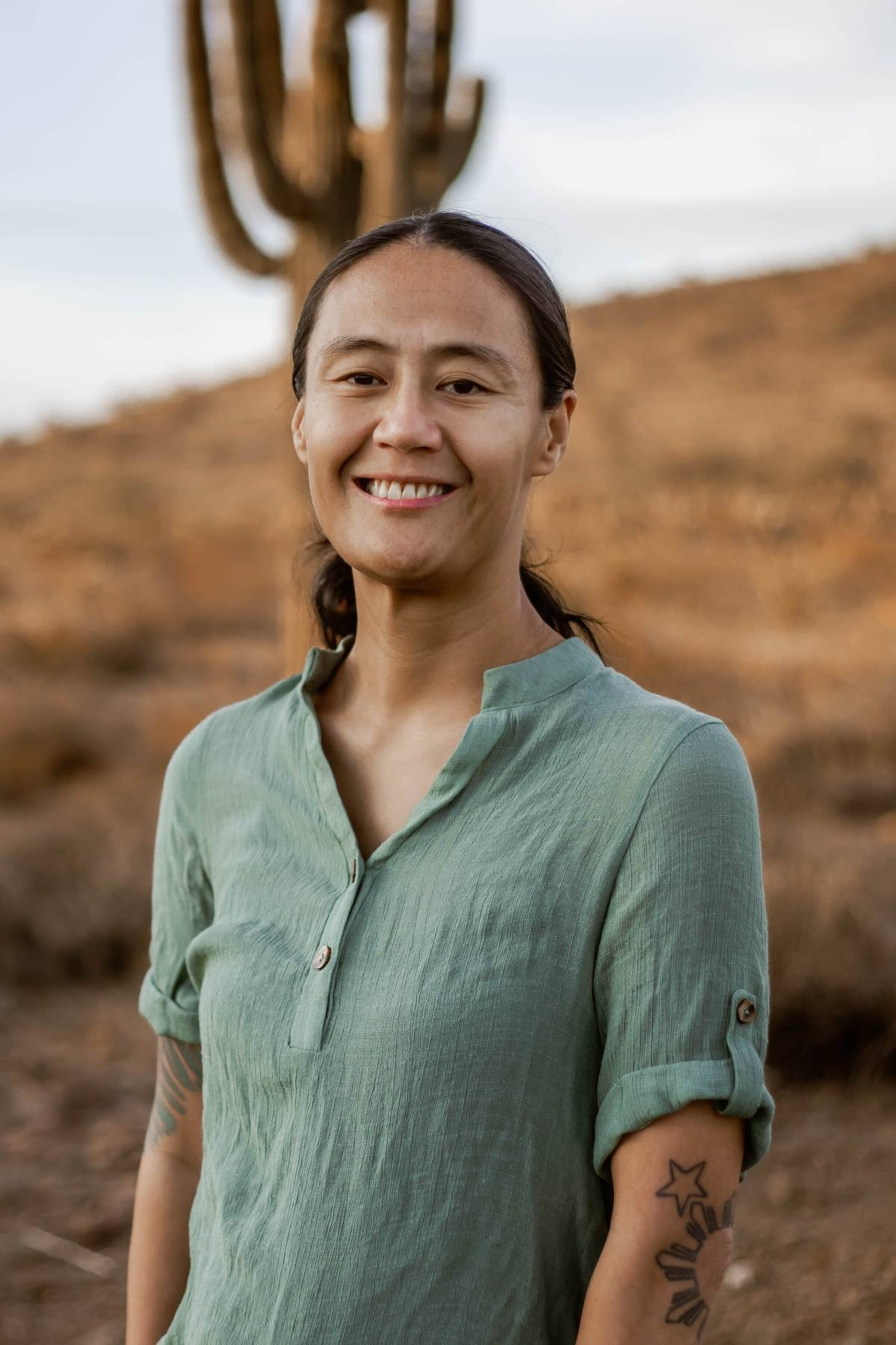Mei Nishimoto, an Asian tattooed brunette in a green top in front of a desert landscape. Photo credit: Jessica Madsen Photography.